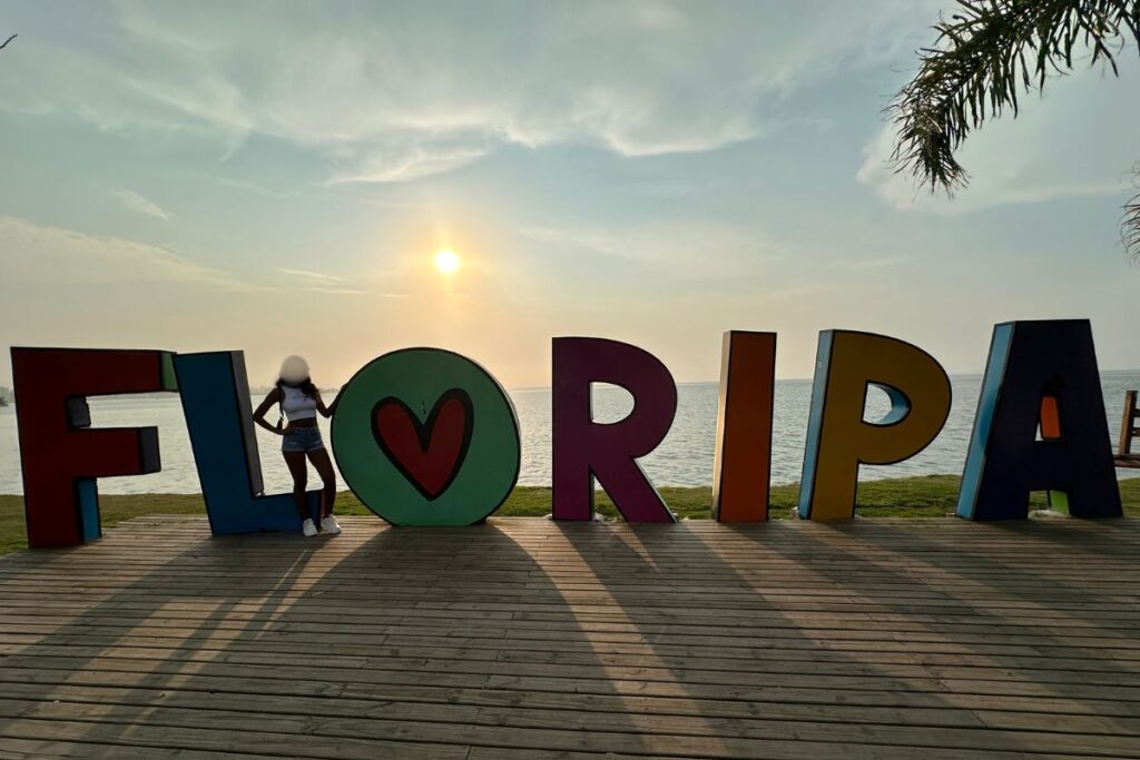 Floripa sign in Florianopolis with Brazilian woman standing in front and sunset in background.